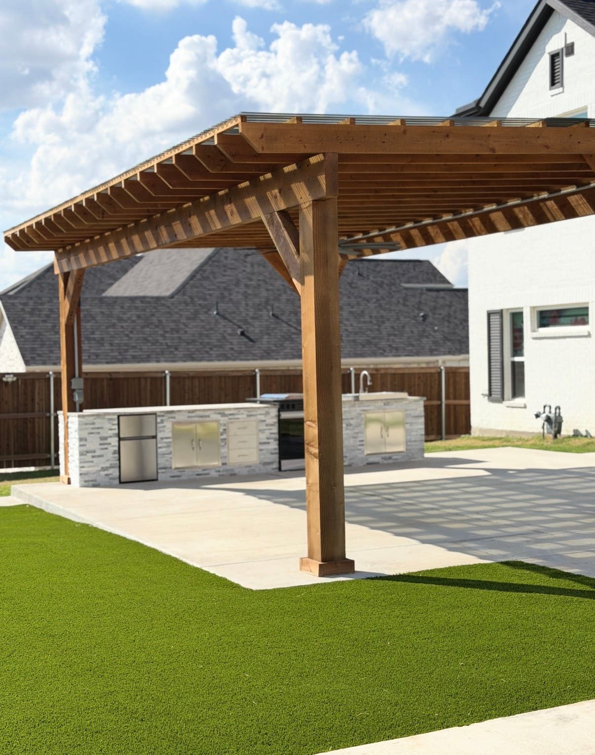Outdoor kitchen with stainless grill, fridge, and sink under a cedar patio cover, with artificial turf in front