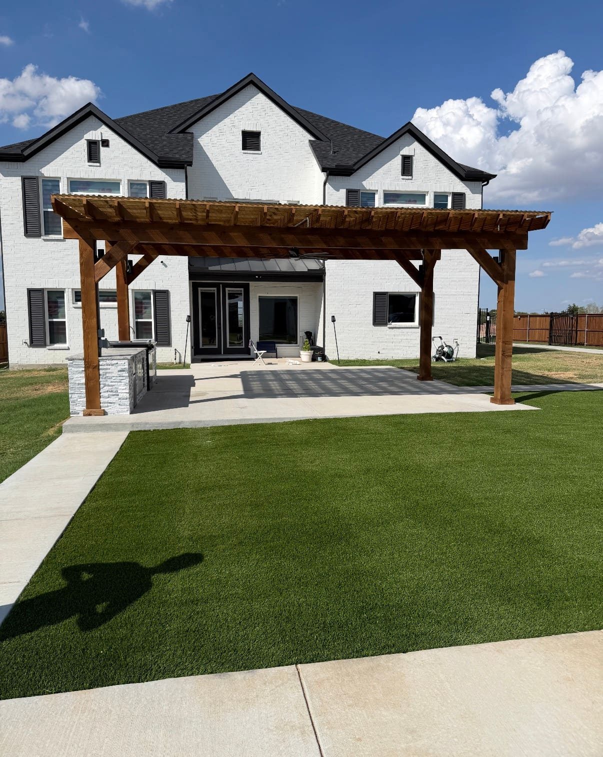 Free-standing cedar patio cover over a concrete patio with an outdoor kitchen and artificial turf behind a white brick home